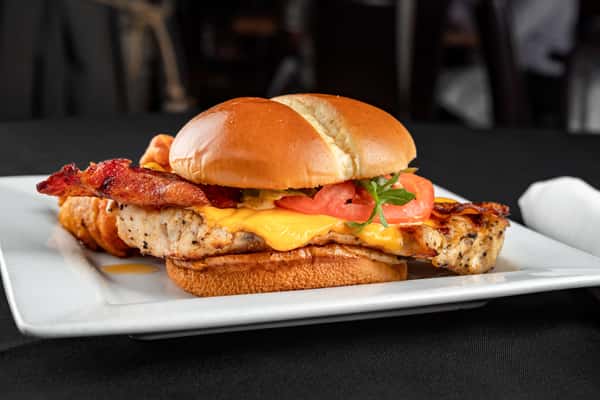 Grilled chicken sandwich with crispy bacon, melted cheddar, fresh tomato, and greens on a toasted brioche bun, paired with seasoned curly fries