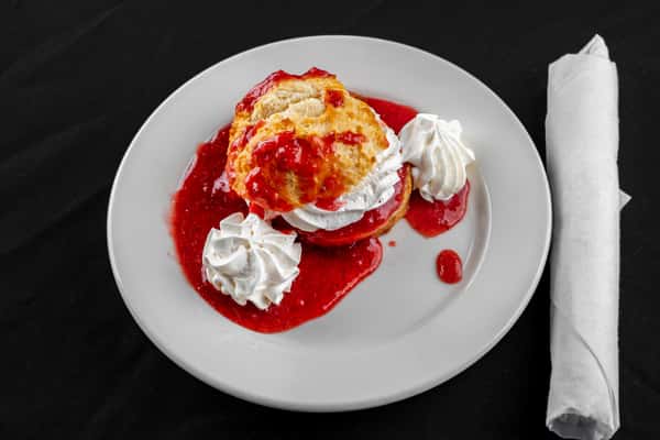 Southern-style strawberry shortcake featuring a fluffy biscuit layered with whipped cream and drizzled with strawberry sauce, garnished with whipped topping on a round white plate.