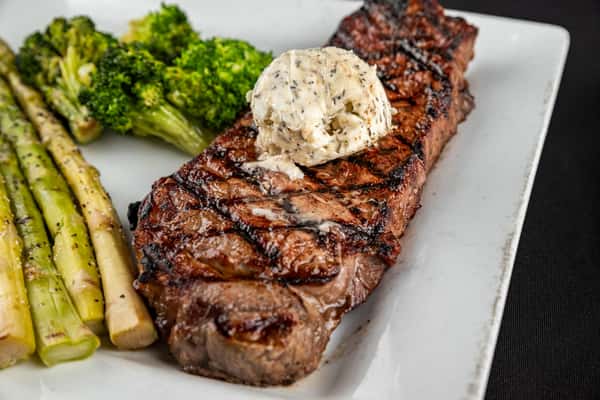 Juicy grilled New York strip steak topped with a dollop of herb butter, served with seasoned asparagus spears and steamed broccoli on a white plate.