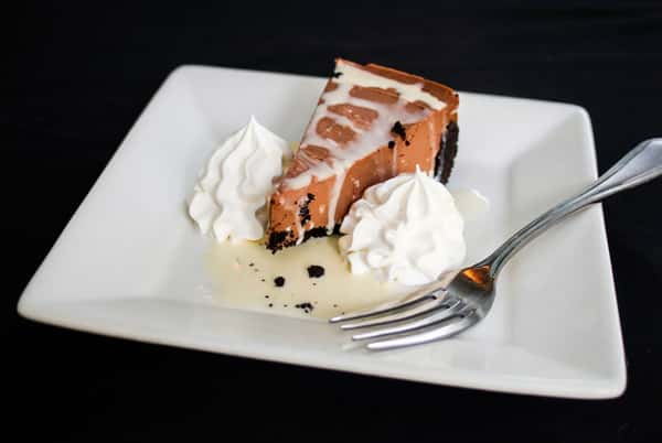 Slice of chocolate cake with white frosting drizzle, served with whipped cream and a fork on a white plate.