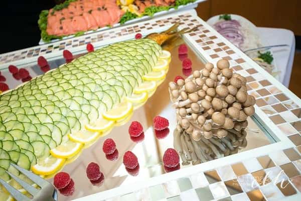 decorative catering tray with a whole smoked fish topped with thinly sliced cucumbers, lined with lemon slices. on a tray with a bundle of raw mushrooms and raspberries for garnish
