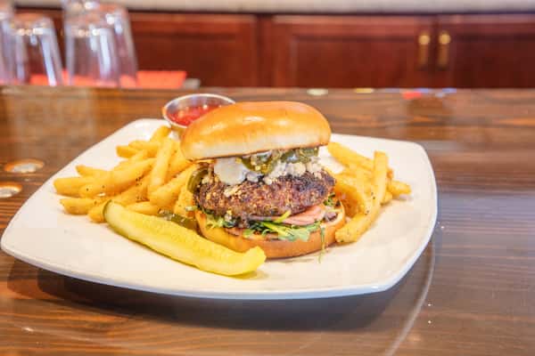 Gourmet burger with blue cheese crumbles and jalapeños on a toasted bun, served with seasoned fries and a pickle on a bar counter