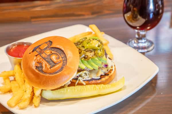 Bacon cheeseburger with crispy onion straws and BBQ sauce on a branded brioche bun, served with seasoned fries and a pickle at a craft brewery restaurant