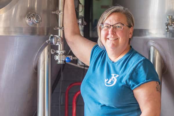 Smiling brewery staff member standing beside stainless steel brewing tanks inside a craft brewery production space