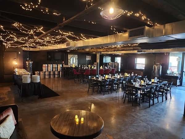 Spacious brewery interior with string lights hanging from the ceiling, dining tables arranged for guests, a central bar area with red stools, and a buffet table set with plates; beer kegs are stacked along the wall.