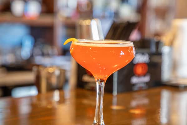 Bright orange craft cocktail in a coupe glass with citrus garnish on a bar counter in a modern brewery