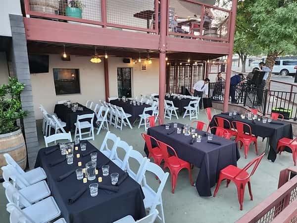 Outdoor dining patio with black tablecloths, red and white chairs, and string lighting under a two-story wooden structure.