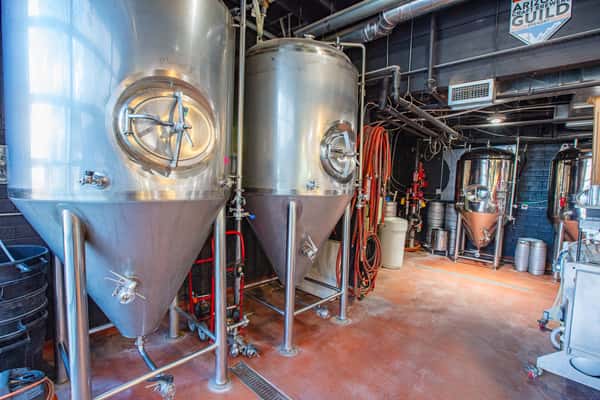Stainless steel fermentation tanks inside a craft brewery production area with industrial brewing equipment and piping visible