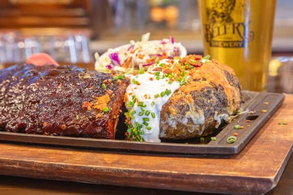 BBQ ribs platter with baked potato and creamy coleslaw served on a wooden board at a craft brewery restaurant with bar taps in the background
