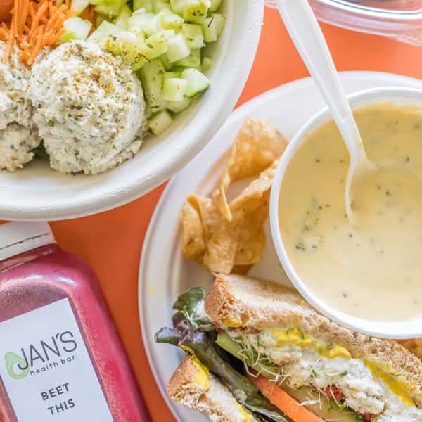 A group photo of a Protein Bowl with Chicken Salad, Cucumber, & Carrots; a Surf Shop Special with half of a Jan's Classic Chicken Salad Sandwich, a cup of Broccoli Cheese Soup, and Have'A Corn Chips; and a bottle of Beet This Juice.