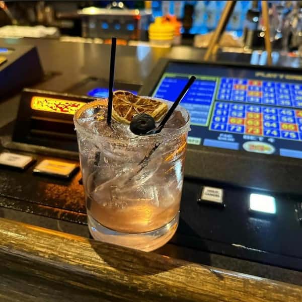 A pale cocktail over ice, garnished with dried citrus and a dark cherry at the bar.