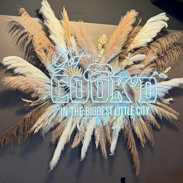 A glowing "Get Cook'd in the Biggest Little City" sign surrounded by a dramatic arrangement of pampas grass.