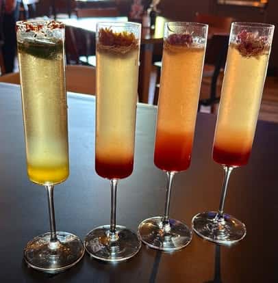 Four layered sparkling cocktails in tall flute glasses, garnished with herbs and dried flowers.