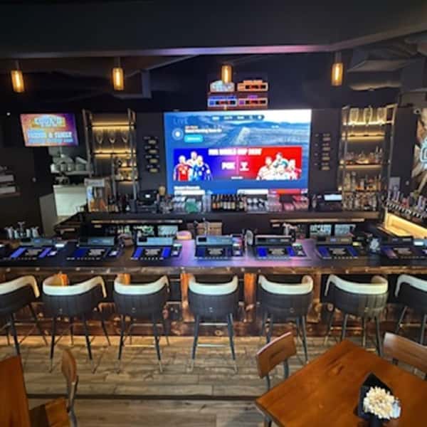 Modern sports bar with counter seating, self-service gaming screens, illuminated bottle displays, and large televisions.