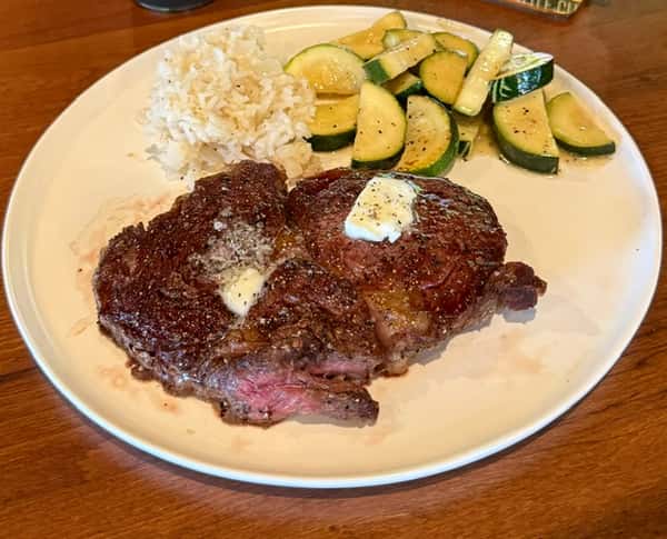 Grilled ribeye steak topped with herb butter, served with sautéed zucchini and rice pilaf