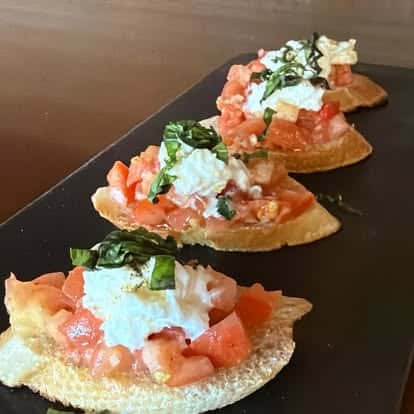 Four tomato-and-cheese crostini topped with fresh basil, arranged on a black serving board.