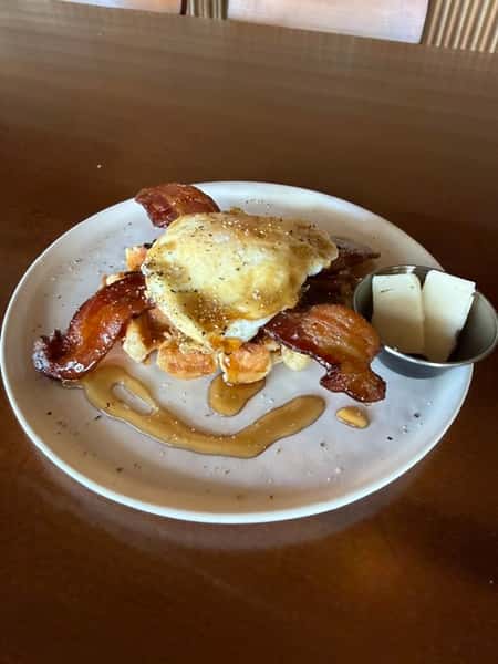 Chicken and waffles topped with a fried egg and crispy bacon, drizzled with syrup, with butter on the side