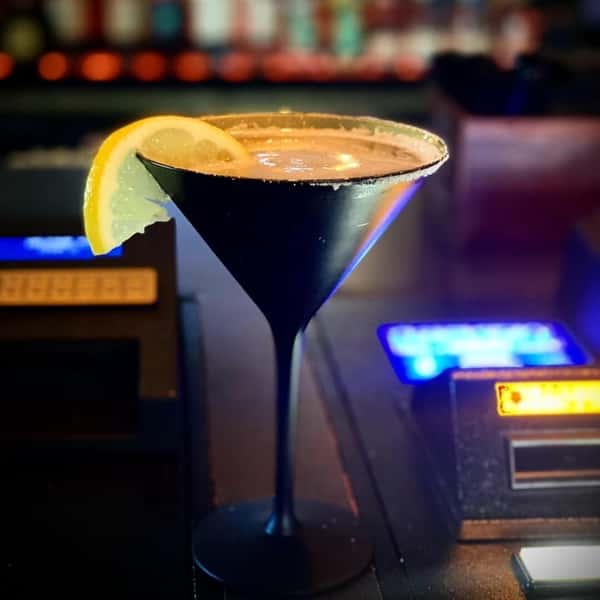A dark cocktail in a martini glass with a sugar-coated rim and lemon-wheel garnish.