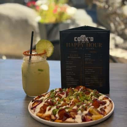 A colorful flatbread pizza and a mason-jar cocktail displayed in front of a Cook'd Happy Hour menu.