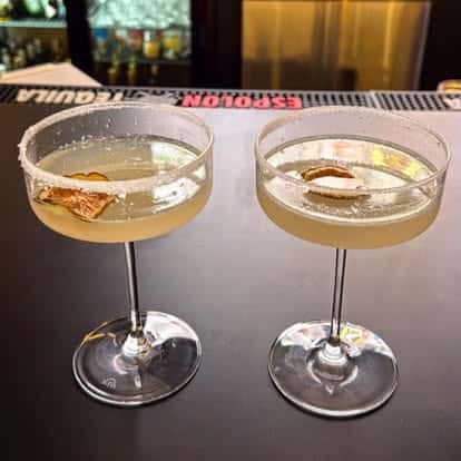 Two pale cocktails in coupe glasses with salted rims and citrus garnishes.