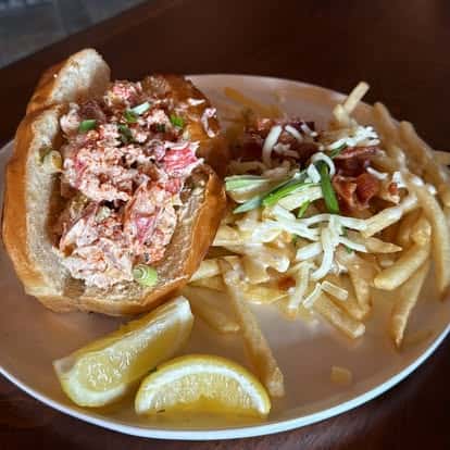 A lobster-roll-style sandwich served with fries, shredded garnish, and two lemon wedges.