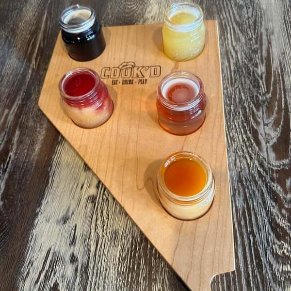Five colorful drink samples in small jars arranged on a wooden Cook'd tasting board.