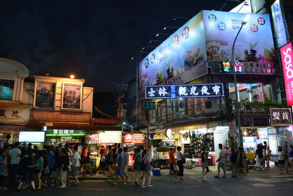 A lively nighttime street scene filled with food stalls, illuminated signs, and visitors at the Shilin Night Market in Taipei, Taiwan.