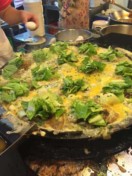 Taiwanese oyster omelets cooking on a griddle with eggs, greens, and chopped scallions
