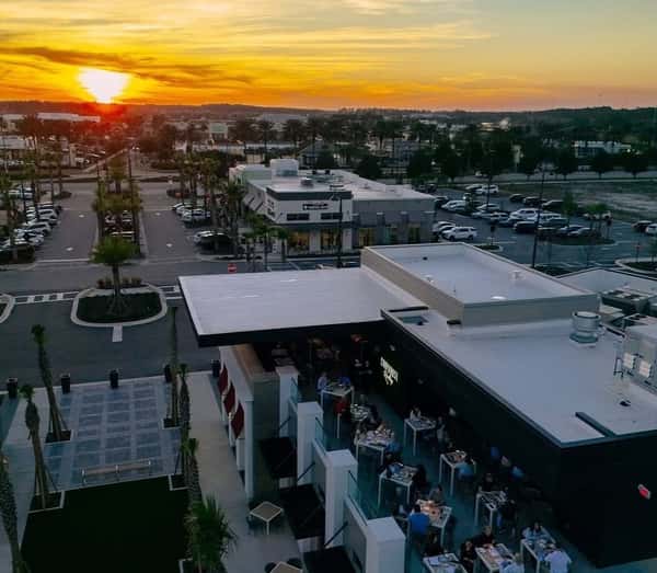 A rooftop restaurant at sunset, with diners seated at tables enjoying the view. The restaurant overlooks a shopping plaza and palm-lined streets, with a vibrant orange and yellow sky in the background.