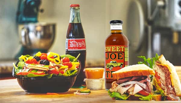 Deli sandwich with salad, dipping sauces, Coca-Cola bottle and Sweet Joe Tea on wooden table