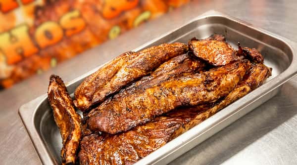 Smoked ribs with a dark, caramelized glaze in a metal tray, with blurry "Ribs & Chops" signage in background.