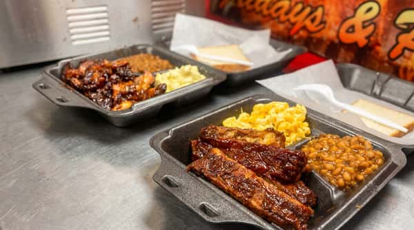 Two take-out containers with BBQ ribs, mac & cheese, and baked beans; one also has chopped brisket.