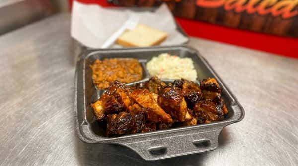 BBQ rib tips with baked beans and coleslaw in a black takeout container, with a slice of white bread.