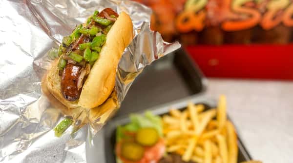 Hot dog in a foil-lined bun with sautéed onions and green peppers, with a blurred burger and fries in background.