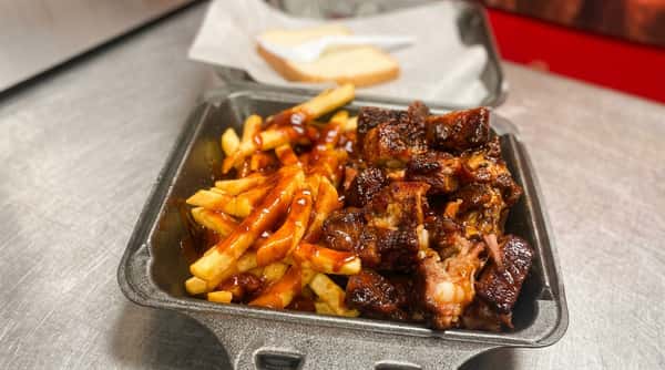 BBQ ribs and french fries covered in sauce in a black to-go container, with two pieces of bread in the background.