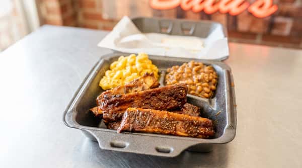 BBQ ribs meal in a black takeout container with macaroni and cheese, baked beans, and a slice of bread.