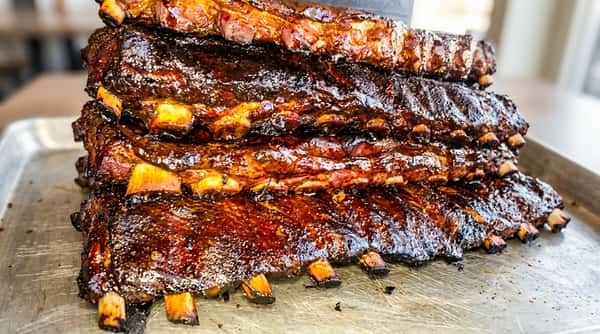 Three racks of smoked BBQ ribs, glistening with a dark, rich sauce, are stacked on a table.