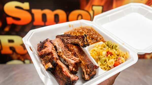 Takeout container with three ribs, baked beans, and pasta salad with tomatoes.