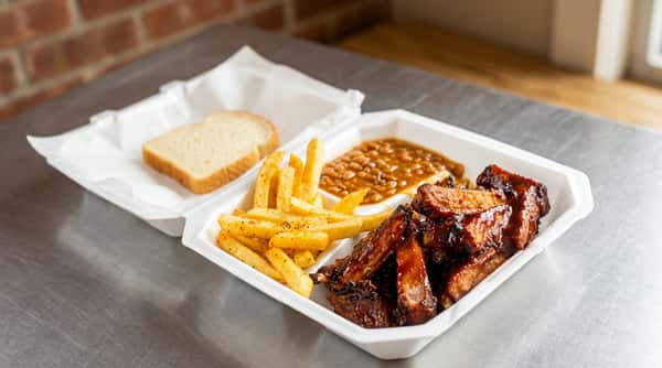 BBQ ribs with fries, baked beans, and white bread in a white takeout container.