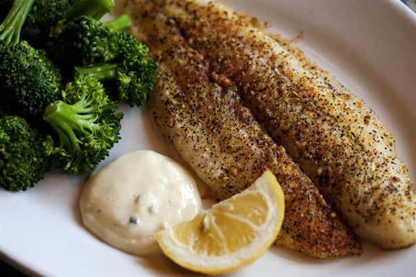 Fish entree cooked on a plate with a small side of broccoli, a lemon wedge and a white creamy sauce.