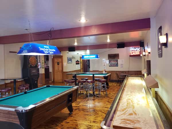 Interior view of The John Hoover Inn featuring green felt pool tables, a shuffleboard, dark wood floors, and beer signage under warm lighting.