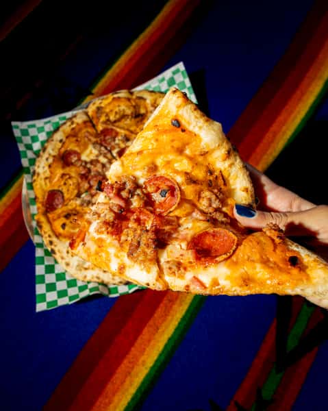Wood-fired pizza topped with pepperoni, sausage, and melted cheese on a blistered crust over colorful textile backdrop