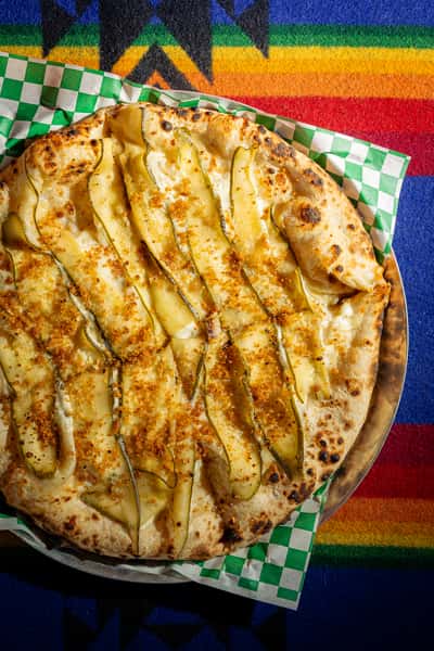 Wood-fired white pizza topped with thinly sliced zucchini, melted cheese, and crispy crust on a colorful textile backdrop