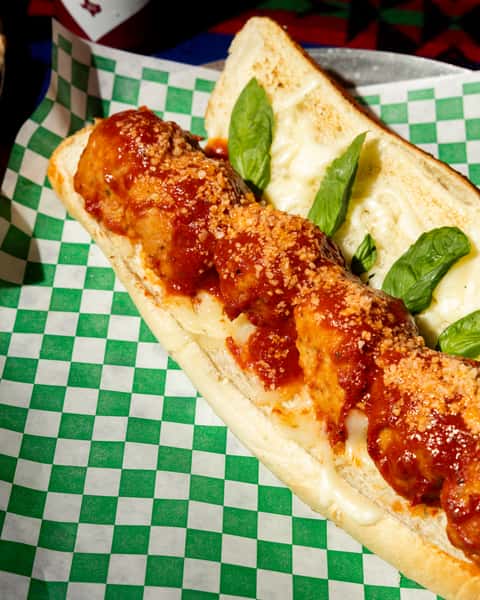 Meatball sub with marinara sauce and grated cheese on toasted bread, garnished with fresh basil on checkered paper.