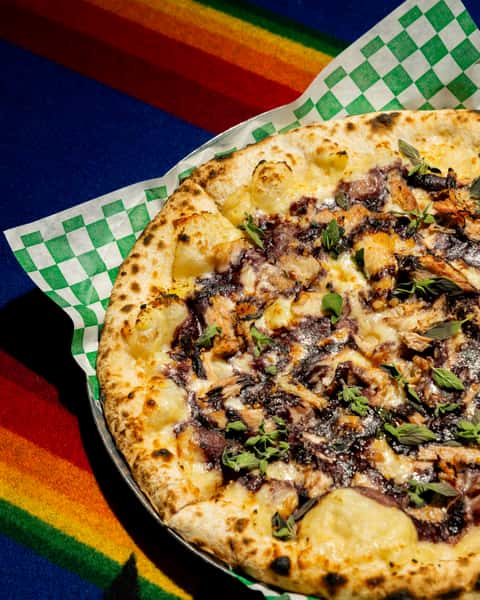 Close-up of wood-fired pizza topped with melted cheese, shredded meat, herbs, and rich dark sauce on blistered crust.