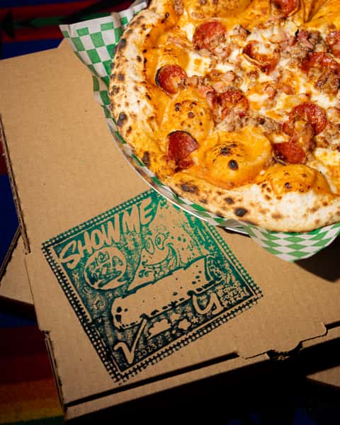 Wood-fired pizza topped with pepperoni, sausage, and melted cheese on a blistered crust over colorful textile backdrop