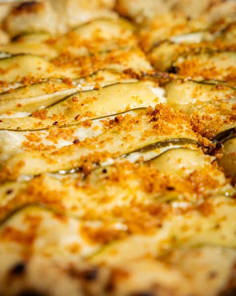 Wood-fired white pizza topped with thinly sliced zucchini, melted cheese, and crispy crust on a colorful textile backdrop