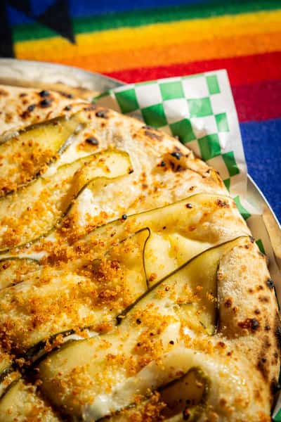 Wood-fired white pizza topped with thinly sliced zucchini, melted cheese, and crispy crust on a colorful textile backdrop