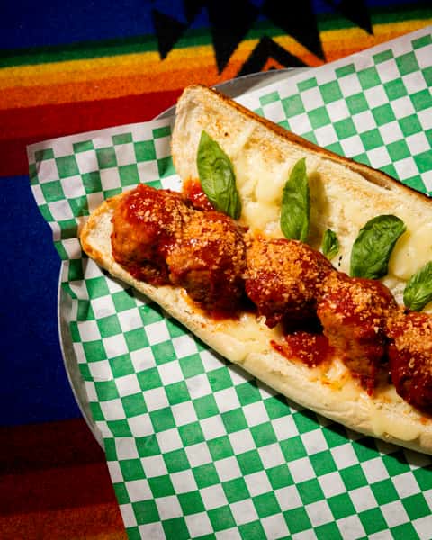 Meatball sub with marinara sauce and grated cheese on toasted bread, garnished with fresh basil on checkered paper.