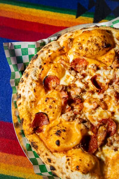 Wood-fired pizza topped with pepperoni, sausage, and melted cheese on a blistered crust over colorful textile backdrop